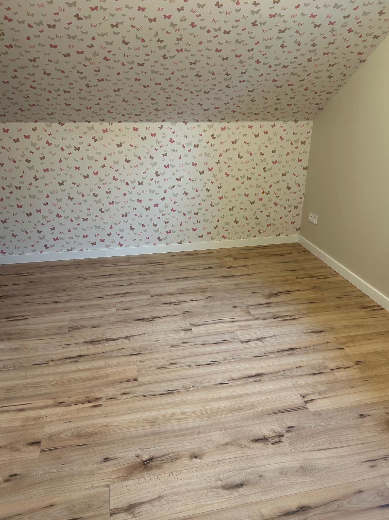 Jasne panele podłogowe w pokoju na poddaszu z tapetą w motyle. Widoczny fragment ściany w kolorze beżowym i biała listwa przypodłogowa.