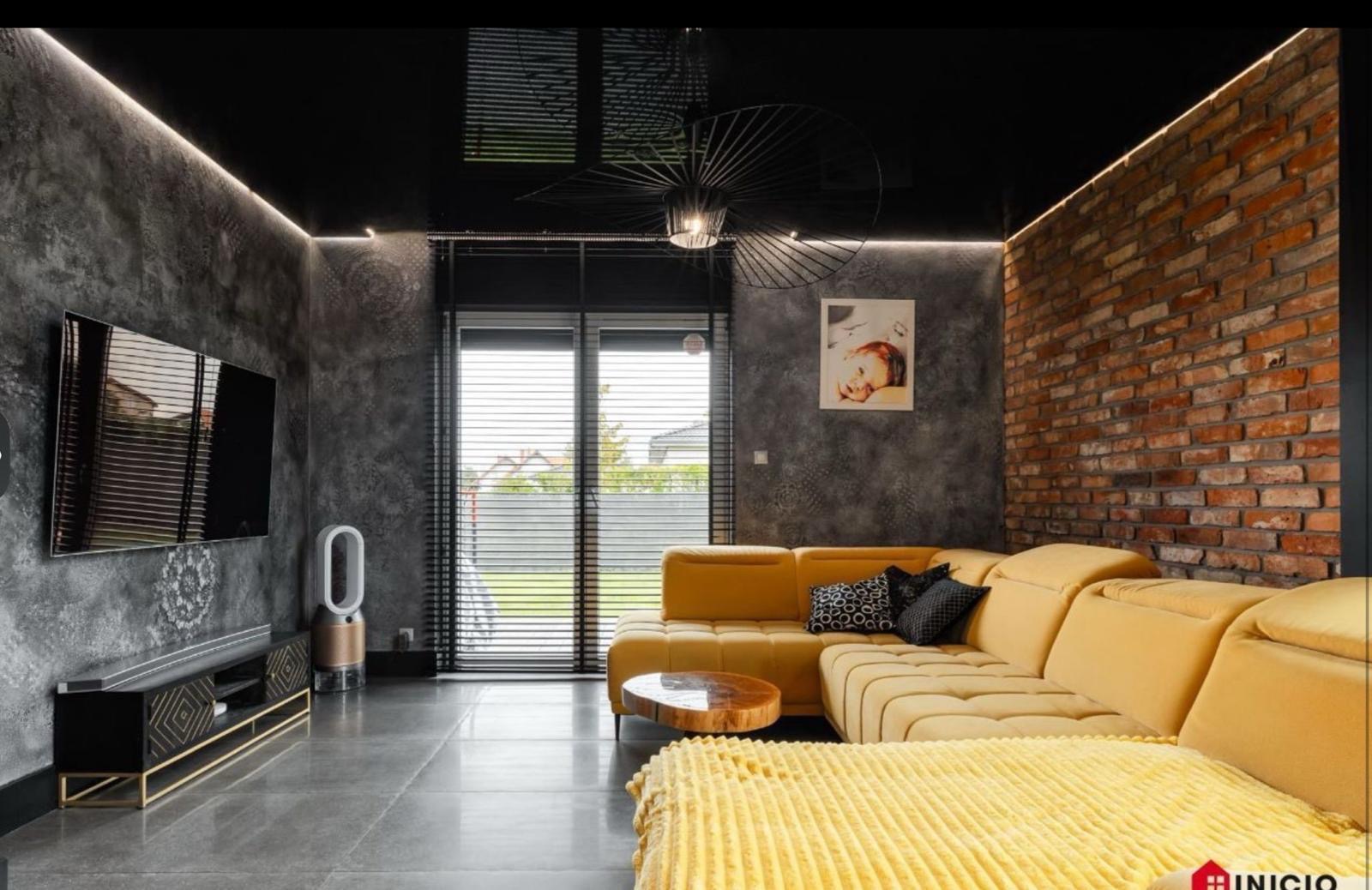 Nowoczesny salon z szarą ścianą o dekoracyjnej fakturze, ceglaną ścianą, żółtą sofą narożną, błyszczącym czarnym sufitem i designerską lampą sufitową.