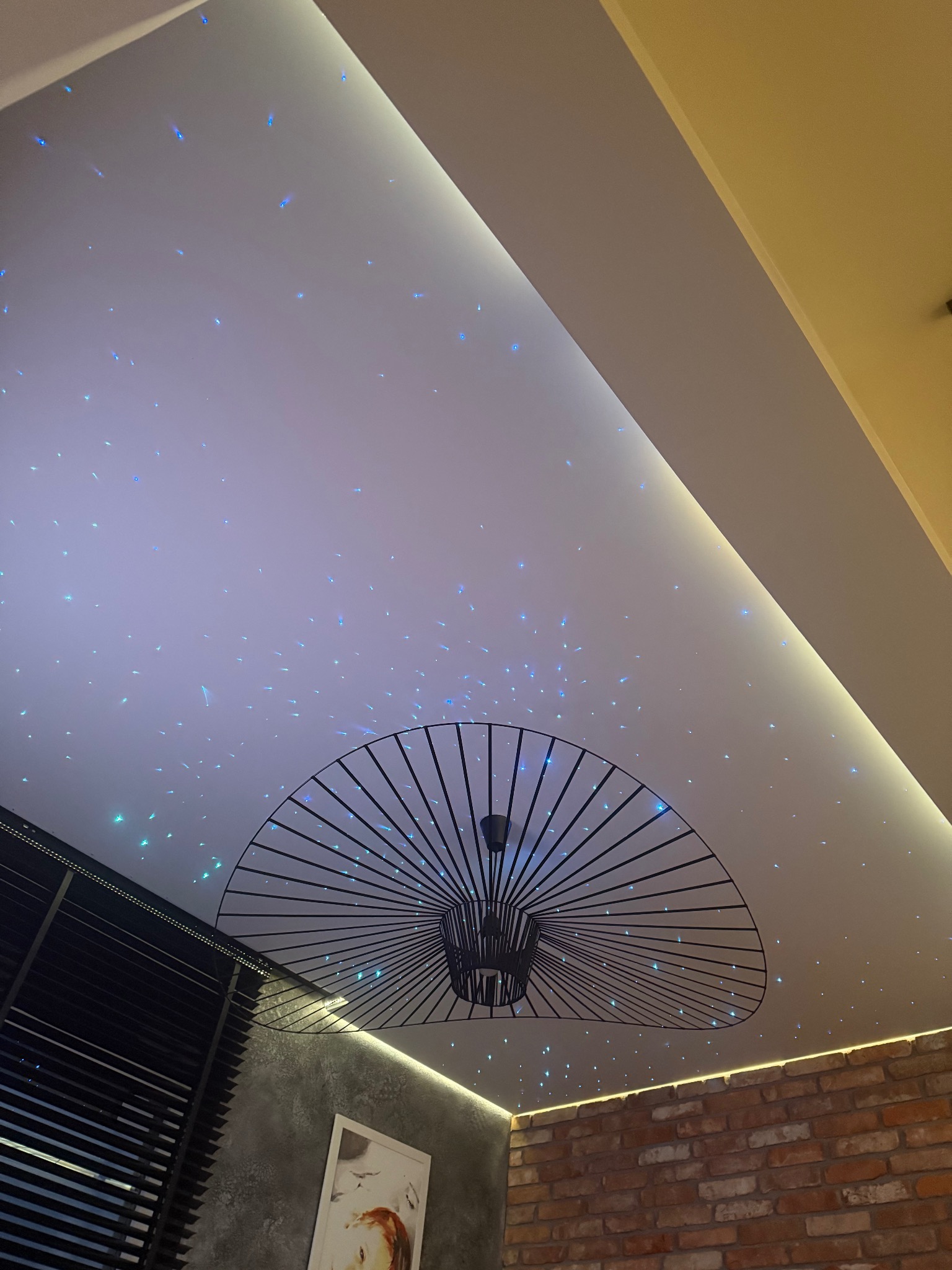 Biały sufit napinany z efektem rozgwieżdżonego nieba, oświetlony listwą LED, z nowoczesną lampą wiszącą na pierwszym planie, widok na fragment ściany z cegły i żaluzje okienne.