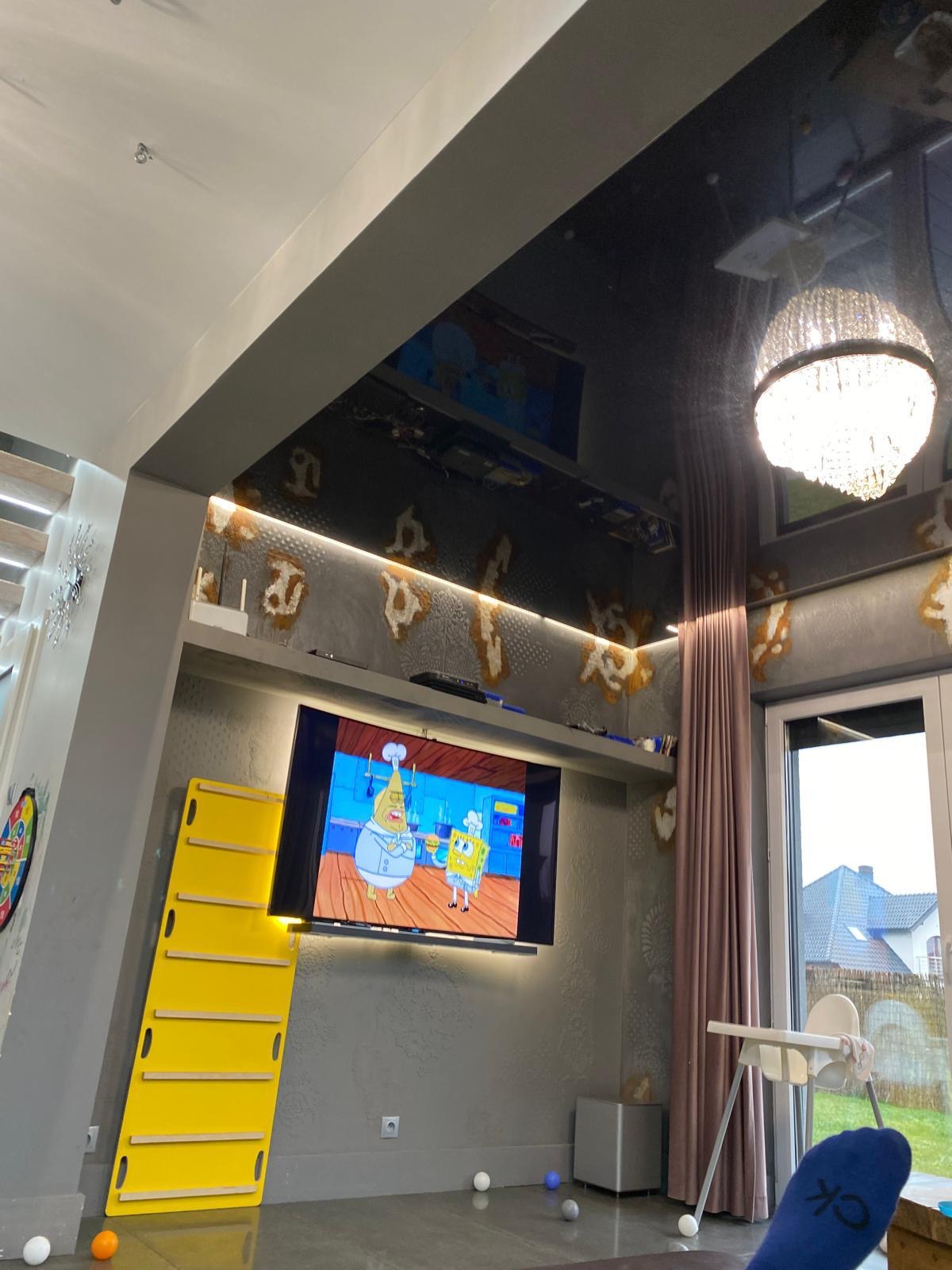 Pokój z błyszczącym, ciemnym sufitem, na którym odbija się żyrandol i telewizor z włączoną bajką, ściany w odcieniach szarości z dekoracyjnymi elementami, żółta drabinka oparta o ścianę.