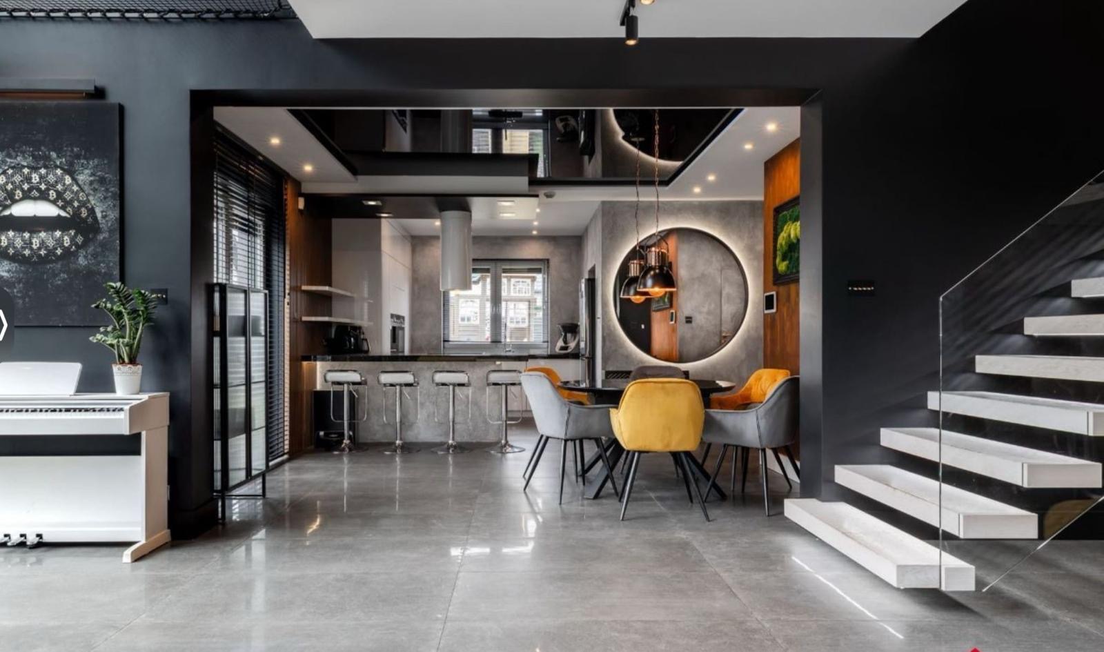 Nowoczesne wnętrze domu z szarą posadzką, widok na otwartą kuchnię z barkiem, jadalnię z okrągłym lustrem i schody z podświetleniem.