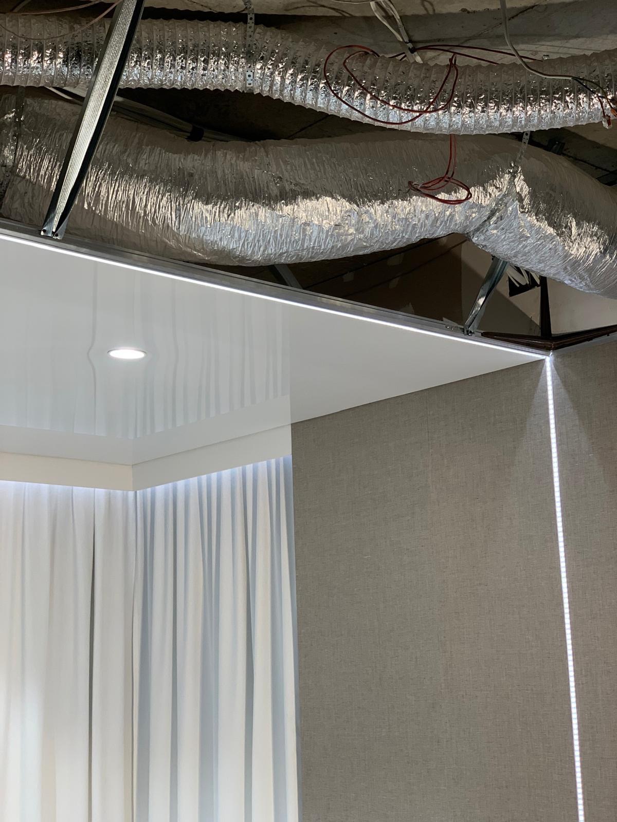 Fragment wnętrza w trakcie prac wykończeniowych: widoczny fragment sufitu z instalacją wentylacyjną, białe zasłony oraz szara ściana z oświetleniem LED w narożniku.