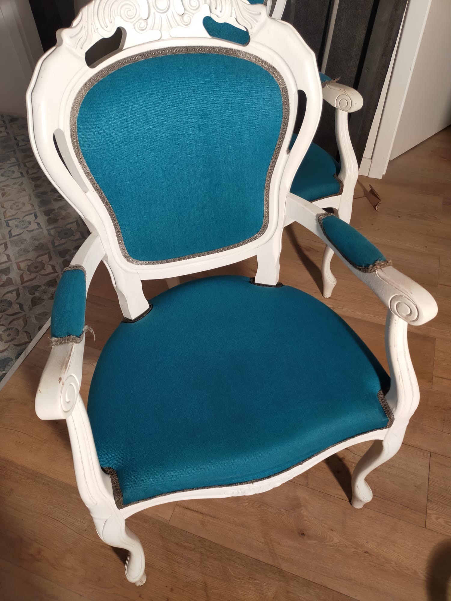 Białe krzesło w stylu Ludwika z turkusową tapicerką, widoczne ślady użytkowania na podłokietnikach, w tle fragment drugiego krzesła i podłoga z wzorem.