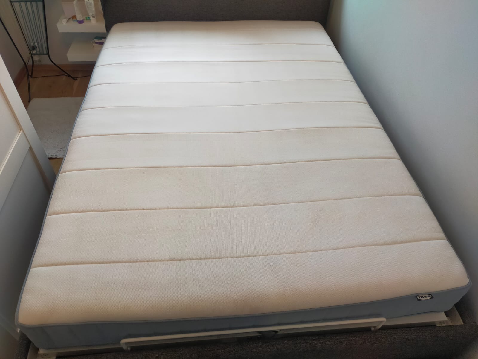 Beżowy materac IKEA z poziomymi przeszyciami, leżący na szarym łóżku, widoczny fragment białej ściany i półki.