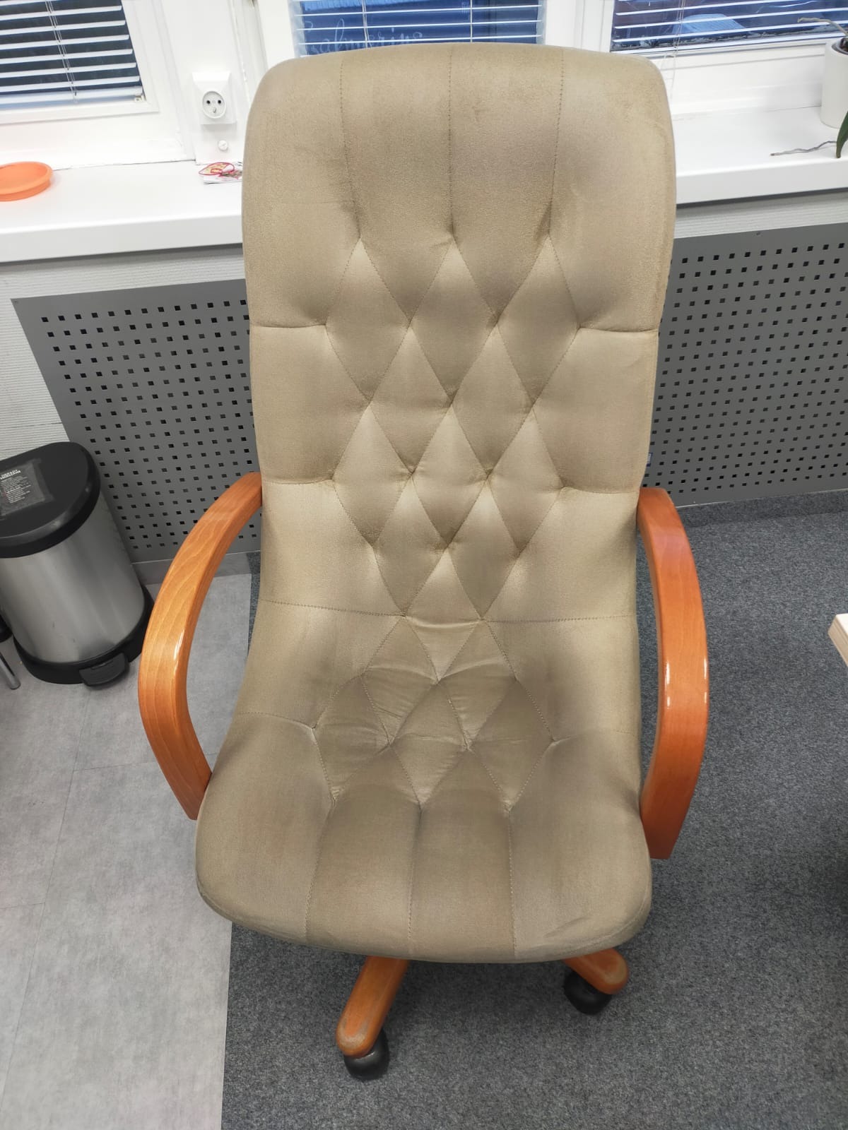 Beżowe krzesło biurowe z drewnianymi podłokietnikami i pikowanym oparciem, widoczne z góry, obok kosza na śmieci i parapetu z drobnymi przedmiotami.