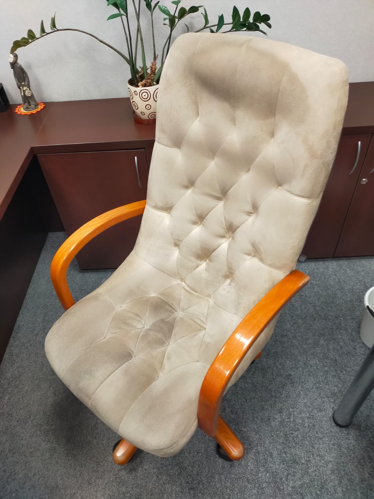 Beżowy fotel biurowy z drewnianymi podłokietnikami, widoczne plamy na tapicerce, na tle biurka i szarej wykładziny.