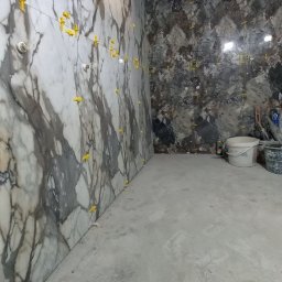 SOFIIA VIKHARIEVA - Wnętrze łazienki w trakcie wykańczania, widoczne płytki imitujące marmur na ścianie z żółtymi krzyżykami dystansowymi, surowy betonowy sufit, narzędzia i wiadra w tle.