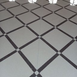 SOFIIA VIKHARIEVA - Ułożona podłoga z jasnych płytek z ciemnymi, ukośnymi paskami tworzącymi wzór kraty; widoczne krzyżyki dystansowe w miejscach łączeń.