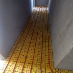 SOFIIA VIKHARIEVA - Instalacja ogrzewania podłogowego z żółtą matą z nadrukowanym wzorem i czerwonymi rurami w surowym, szarym pomieszczeniu podczas remontu.