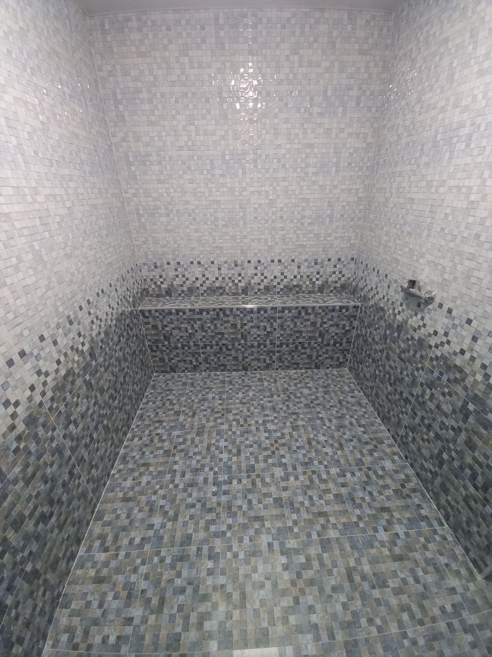 Wykończony prysznic z mozaiką w odcieniach szarości i błękitu, z wnęką na kosmetyki i chromowaną słuchawką prysznicową.