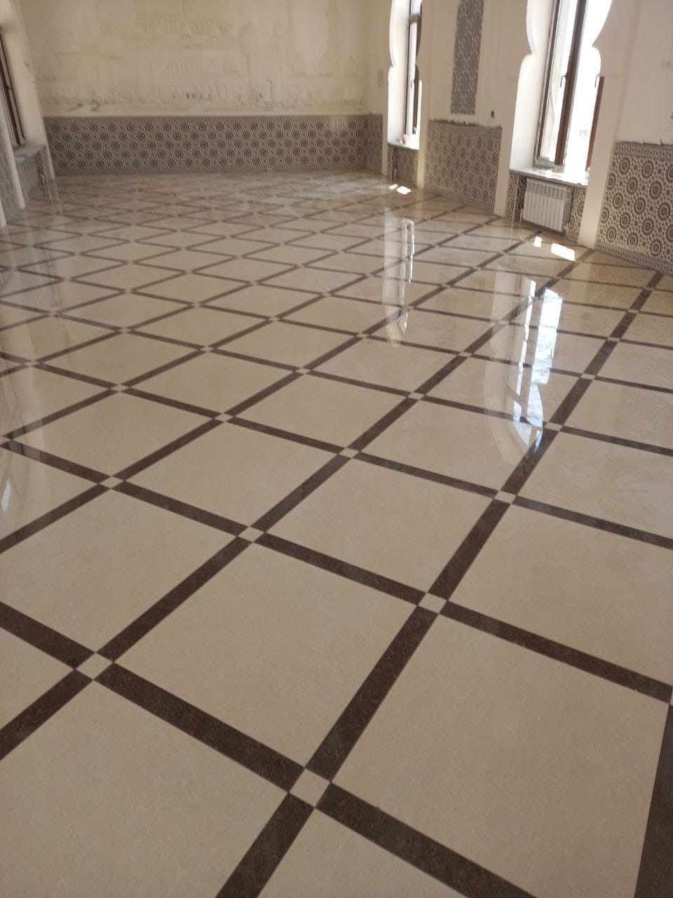 Polerowana podłoga z kwadratowych płytek w beżowo-brązową szachownicę, widoczne fragmenty ścian z dekoracyjnymi płytkami i oknami w pustym pomieszczeniu.