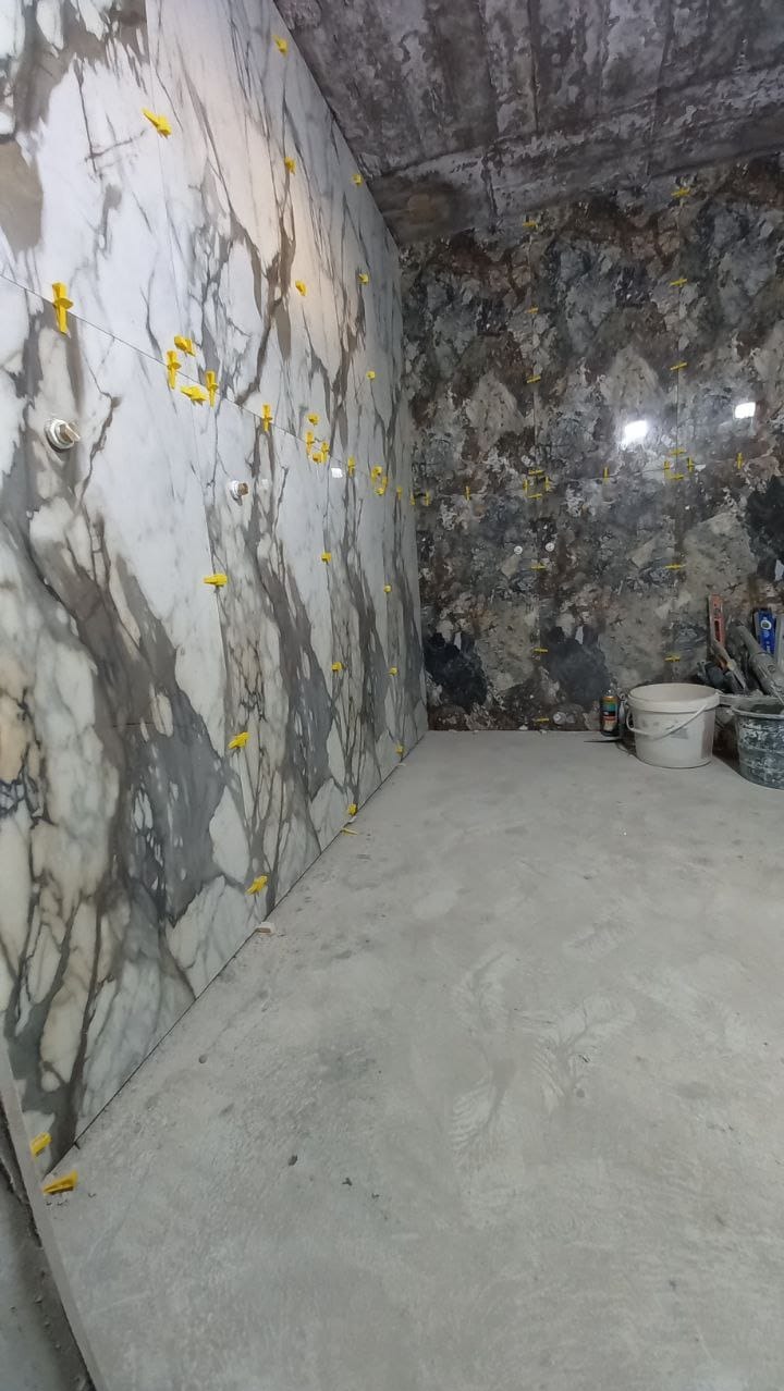 Wnętrze łazienki w trakcie wykańczania, widoczne płytki imitujące marmur na ścianie z żółtymi krzyżykami dystansowymi, surowy betonowy sufit, narzędzia i wiadra w tle.
