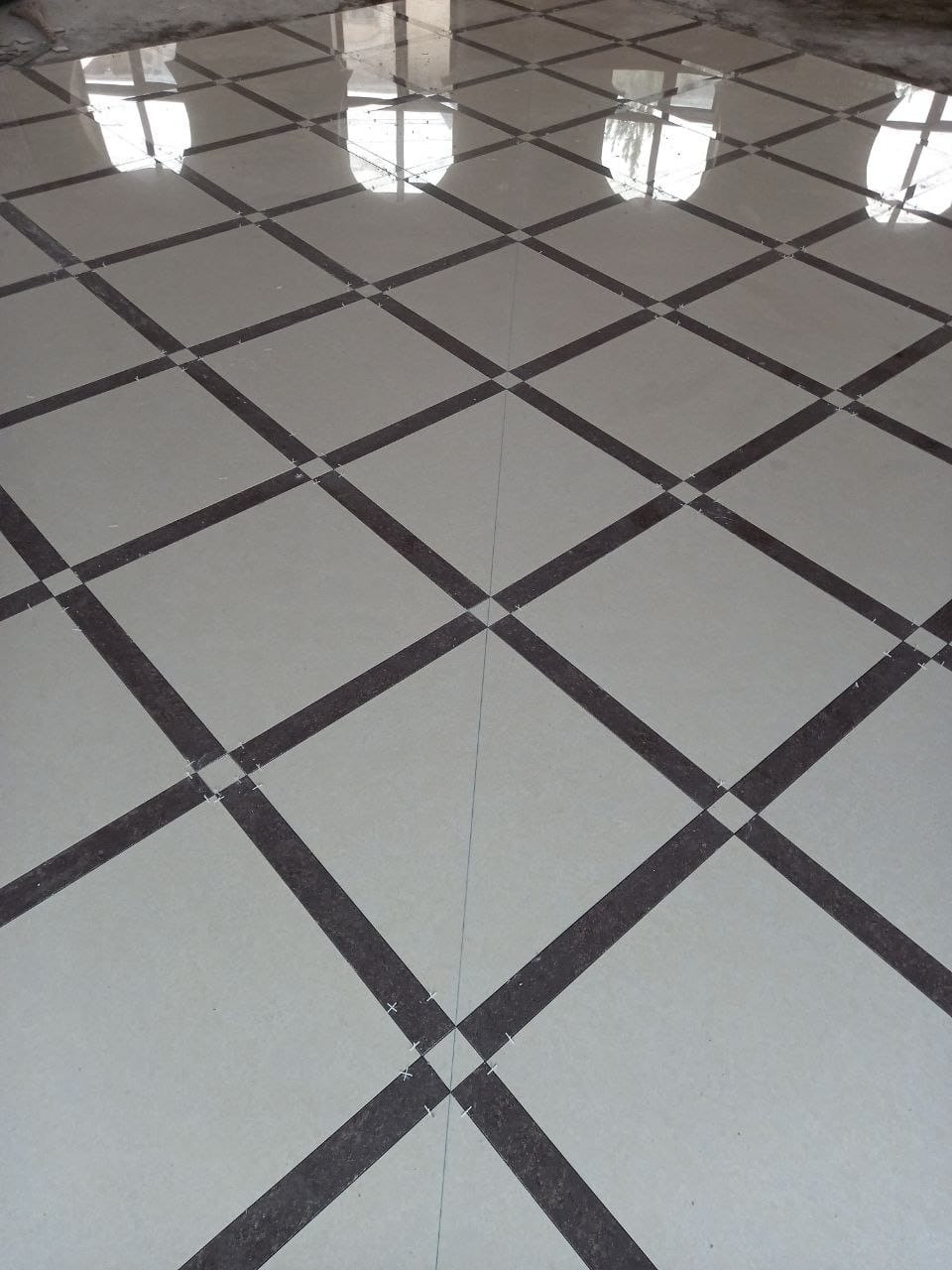 Ułożona podłoga z jasnych płytek z ciemnymi, ukośnymi paskami tworzącymi wzór kraty; widoczne krzyżyki dystansowe w miejscach łączeń.