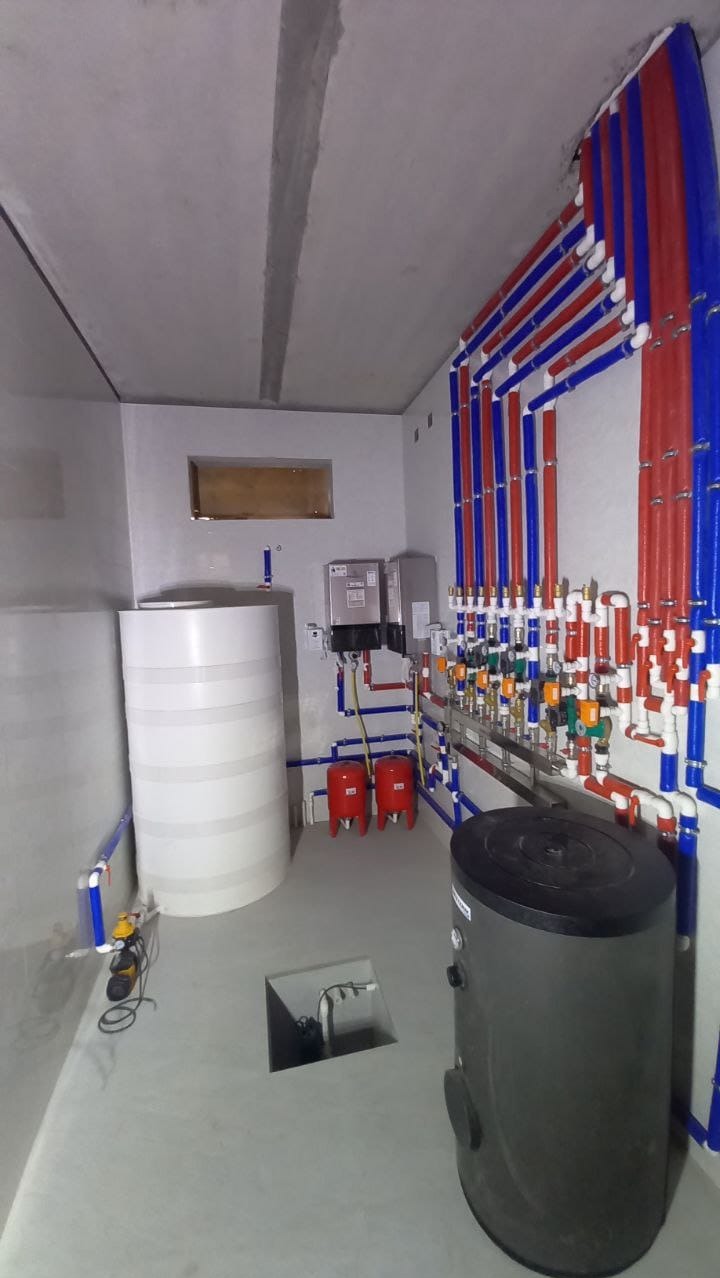 Kotłownia w surowym stanie: widoczne zbiorniki, rury w kolorach czerwonym i niebieskim, instalacja grzewcza oraz narzędzia na podłodze.