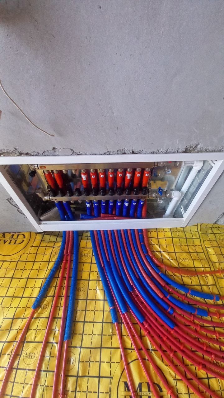 Rozdzielacz ogrzewania podłogowego z czerwonymi i niebieskimi rurkami, zamontowany pod sufitem na żółtej izolacji podłogowej z czarną siatką.