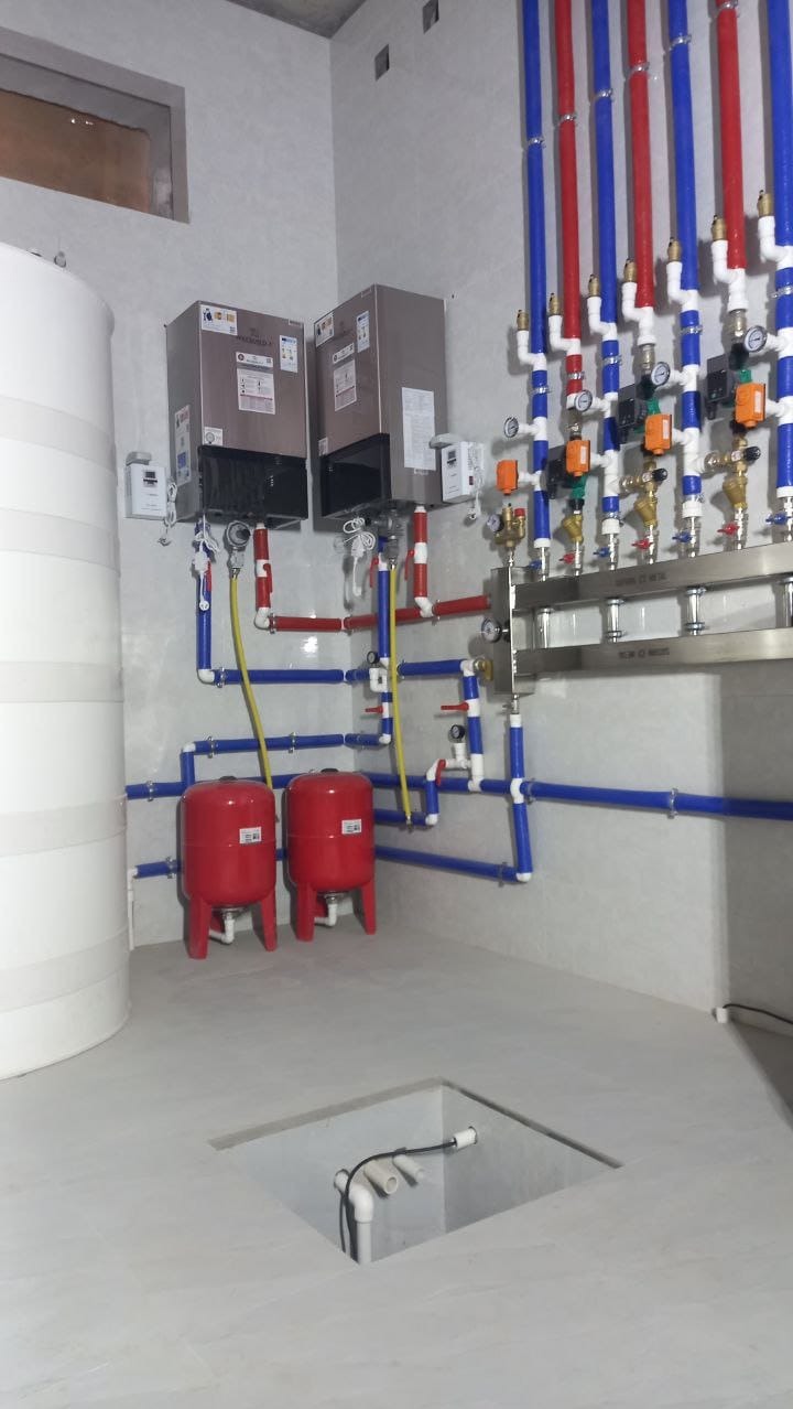 Instalacja centralnego ogrzewania w kotłowni: dwa kotły grzewcze, biały zbiornik akumulacyjny, czerwone zbiorniki przeponowe, rozdzielacz z zaworami, oraz rury instalacyjne w kolorach niebieskim...