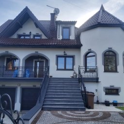 Comfort House s.c. - Danuta Wawrzyńczak, Serhii Pozhyvilko - Dwupiętrowy dom z szarą stolarką okienną, balkonem z kutą balustradą i garażem wbudowanym w bryłę budynku, do którego prowadzą schody z szarej kostki.