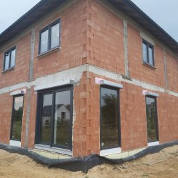 Comfort House s.c. - Danuta Wawrzyńczak, Serhii Pozhyvilko - Surowy stan budynku z cegły z zamontowanymi oknami PCV w grafitowych ramach i częściowo zamontowanymi roletami zewnętrznymi, widoczne ocieplenie i zabezpieczenie fundamentów.