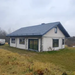 Surowy dom z białego bloczka z zamontowanymi oknami PCV i przesuwnymi drzwiami balkonowymi w czarnych ramach, na tle pochmurnego nieba i jesiennego krajobrazu, z białym busem zaparkowanym obok.