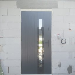 Comfort House s.c. - Danuta Wawrzyńczak, Serhii Pozhyvilko - Szare drzwi zewnętrzne z pionowymi panelami i szybą, zamontowane w surowej ścianie z białych bloczków, widoczne przewody instalacji elektrycznej.