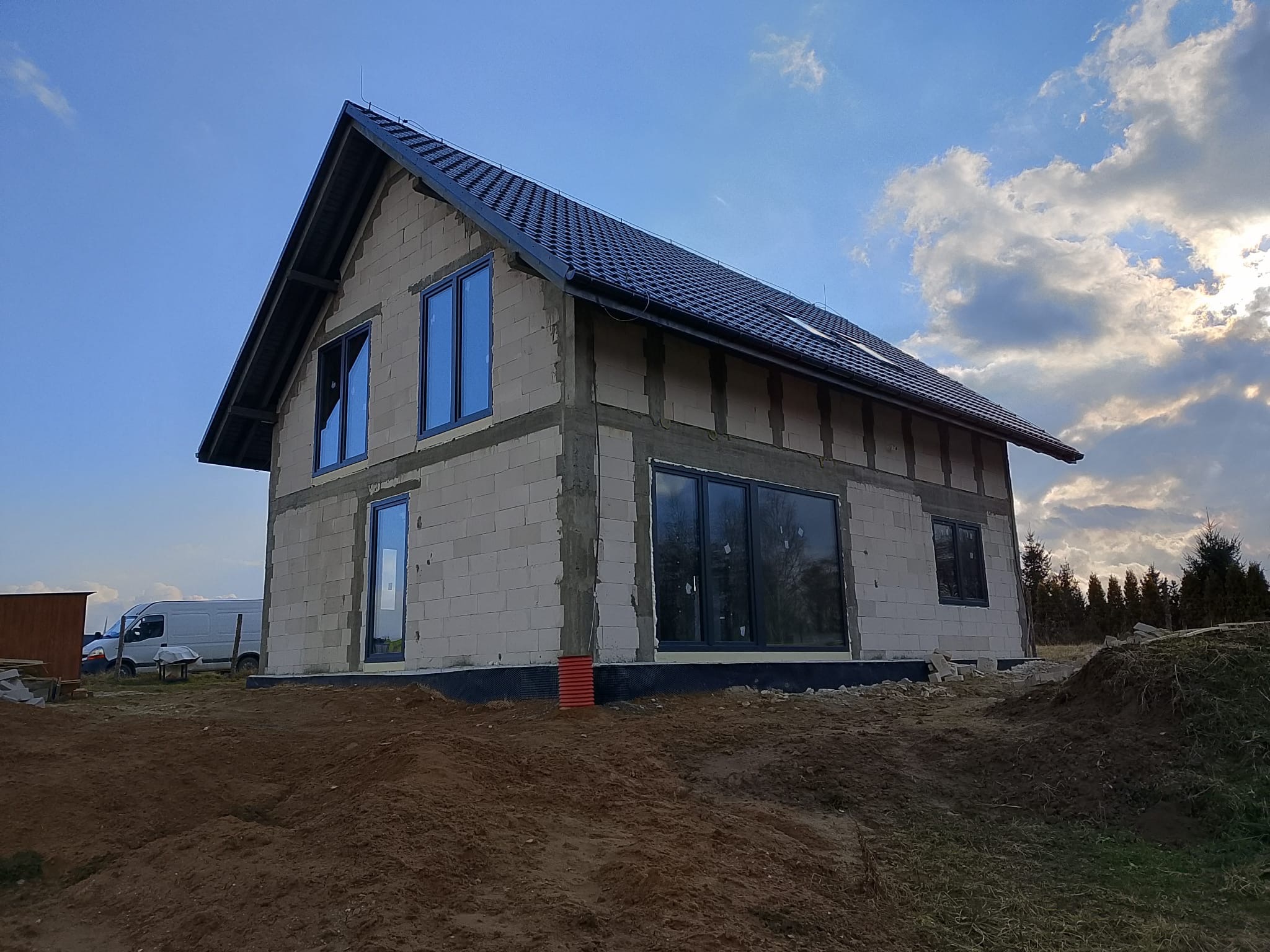 Surowy stan budynku jednorodzinnego z zamontowanymi oknami w grafitowych ramach, widok z zewnątrz na tle zachmurzonego nieba.