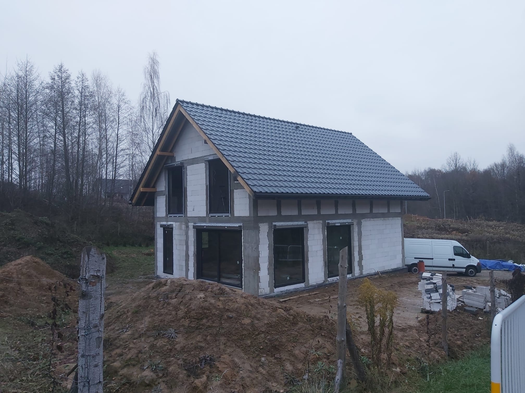 Surowy stan domu w trakcie budowy z zamontowanymi oknami z czarnymi ramami, widoczne bloczki betonowe, dach z ciemnej dachówki, biały samochód dostawczy w tle.