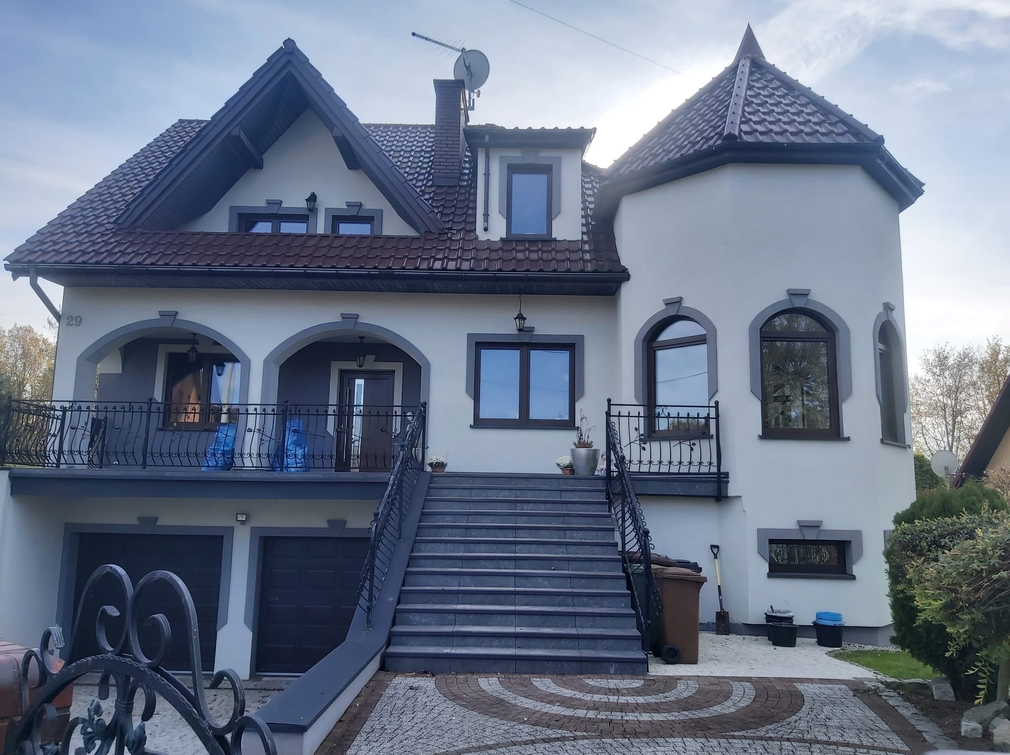 Dwupiętrowy dom z szarą stolarką okienną, balkonem z kutą balustradą i garażem wbudowanym w bryłę budynku, do którego prowadzą schody z szarej kostki.