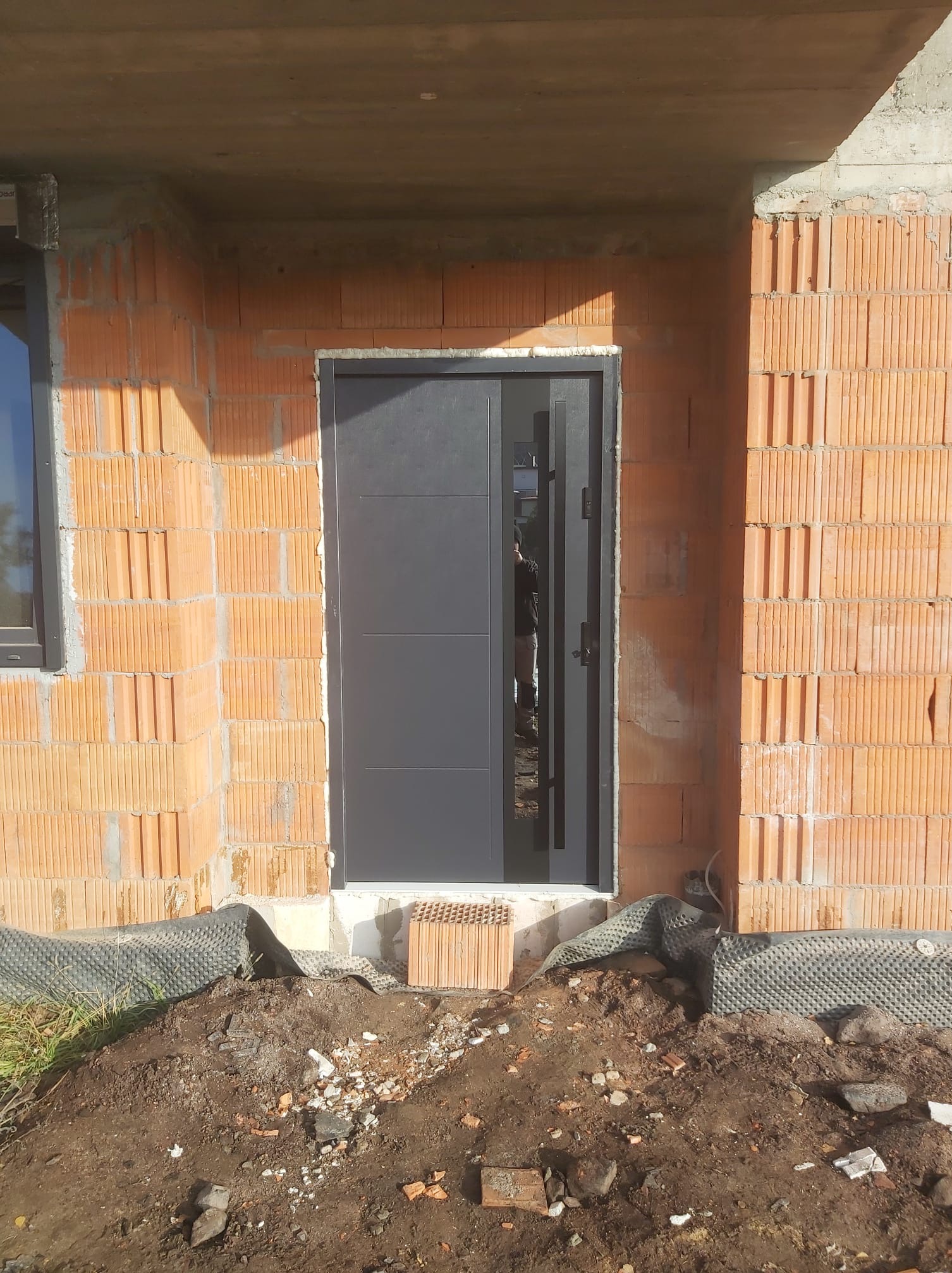 Montaż szarych drzwi wejściowych z bocznym przeszkleniem w surowym murze z czerwonej cegły dziurawki, widoczna izolacja fundamentów i okno w tle.