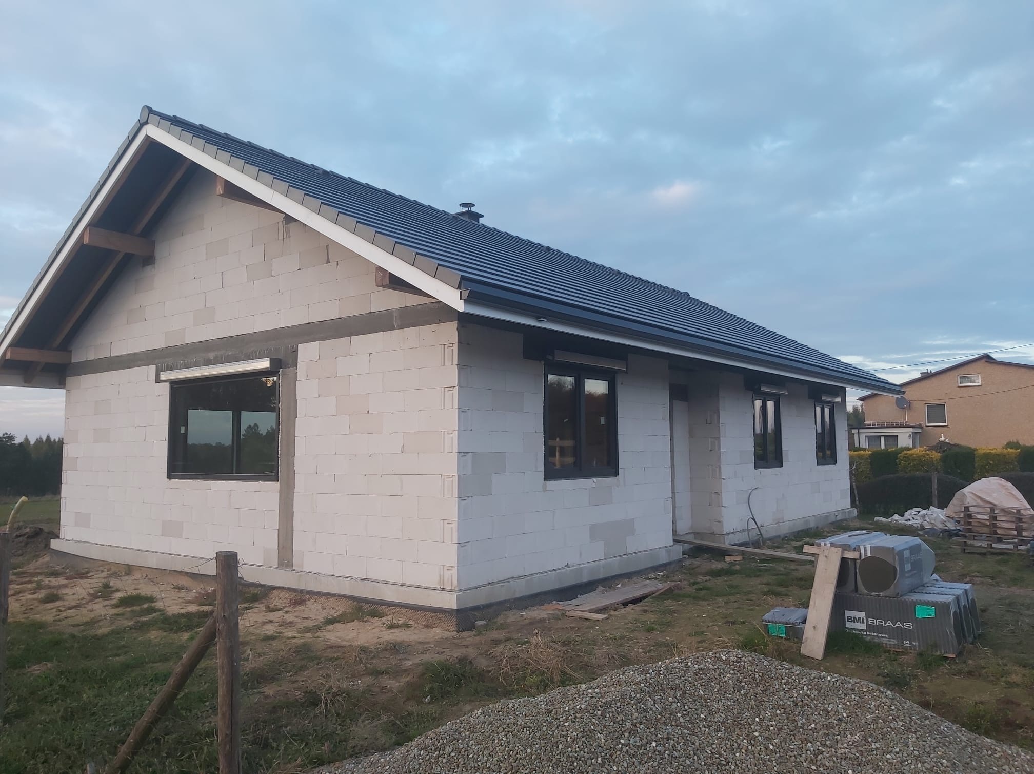 Budowa domu jednorodzinnego z białych bloczków, widoczne czarne ramy okienne z częściowo zamontowanymi roletami zewnętrznymi, niebieska dachówka na dachu, materiały budowlane składowane na posesji.