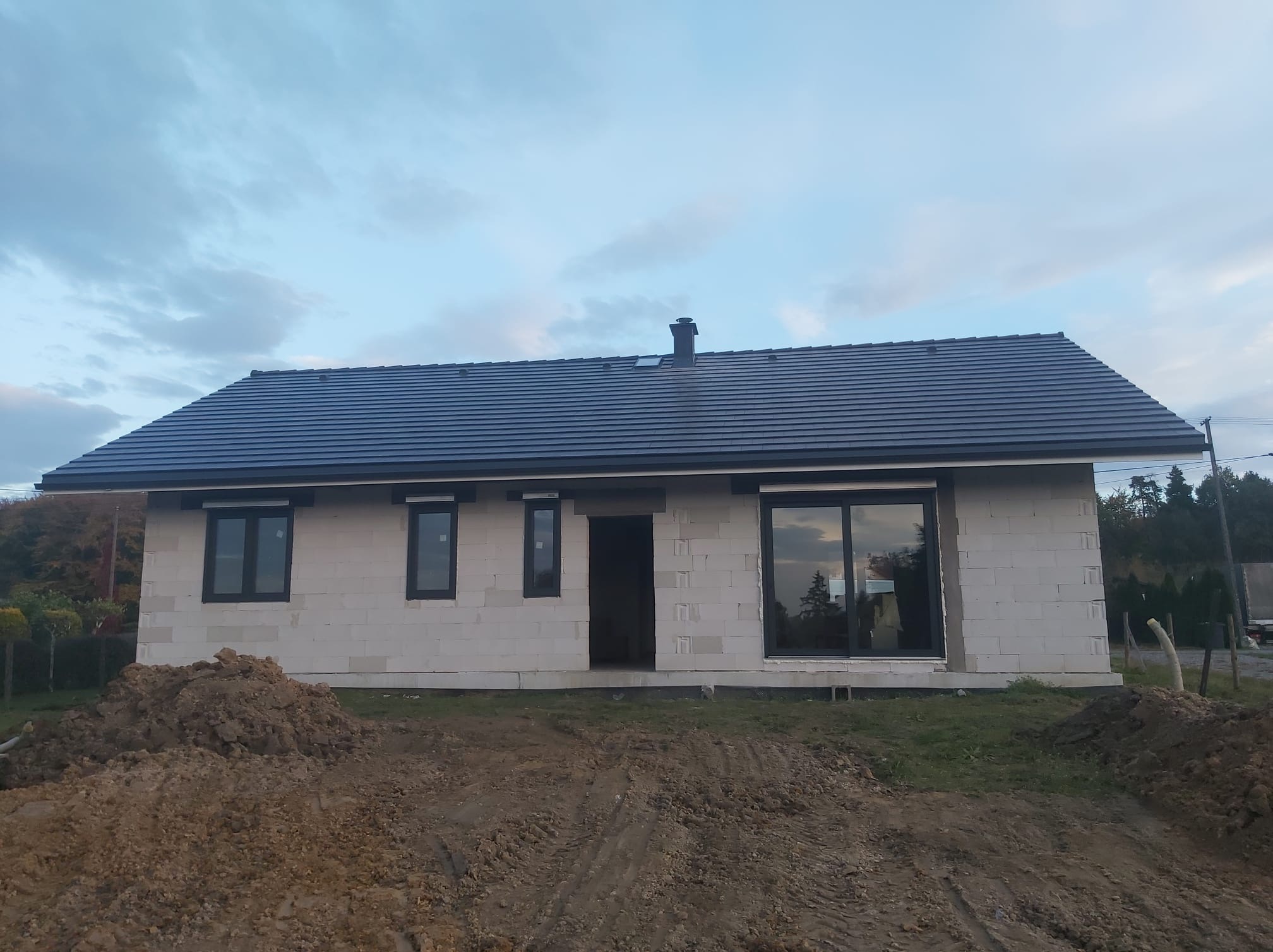 Dom w trakcie budowy z zamontowanymi oknami w czarnych ramach; widoczny stos ziemi na pierwszym planie.
