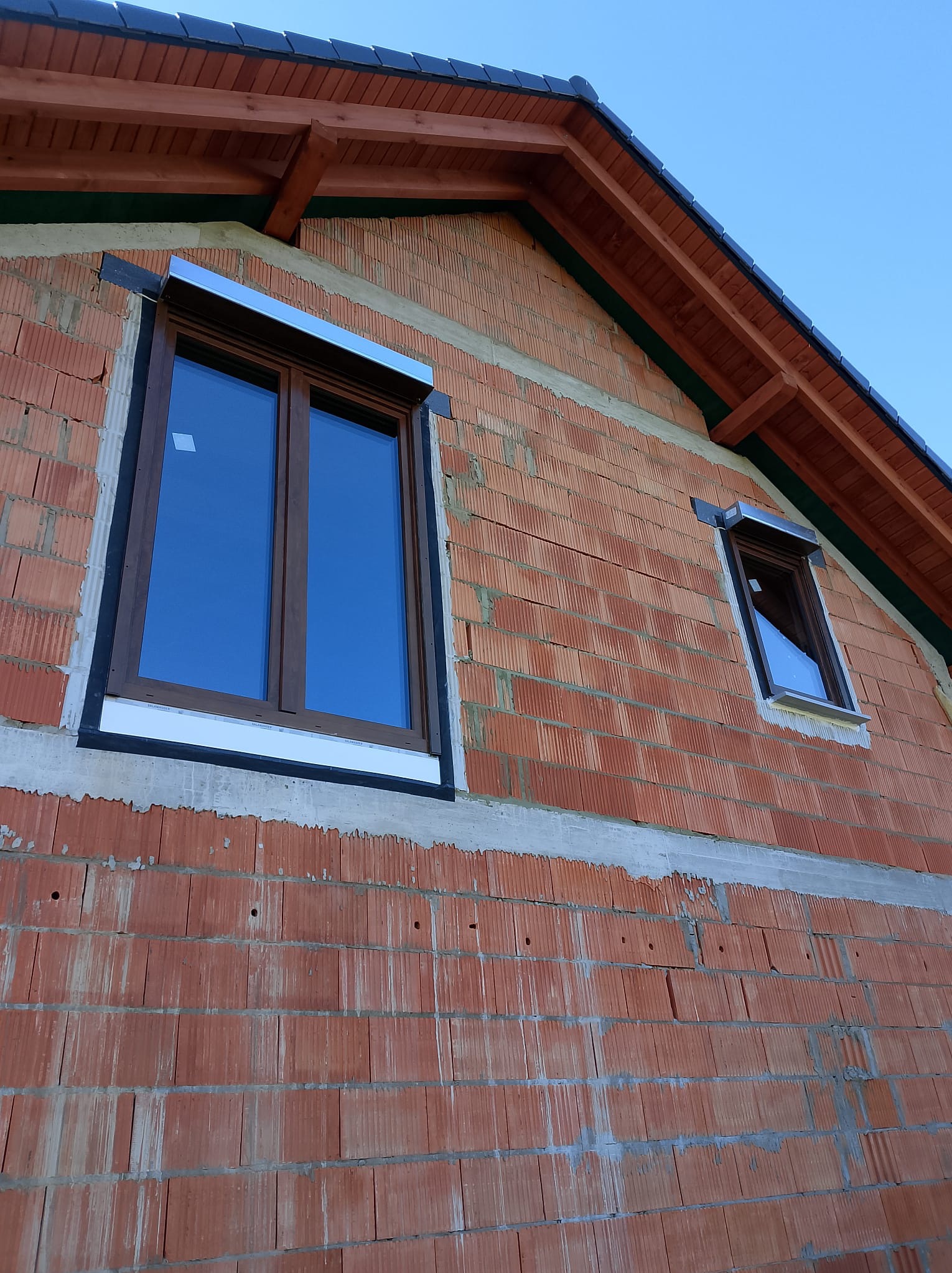 Niedokończona elewacja budynku z czerwonej cegły z zamontowanymi oknami z brązowymi ramami i srebrnymi roletami zewnętrznymi w kasetach, widok z dołu.