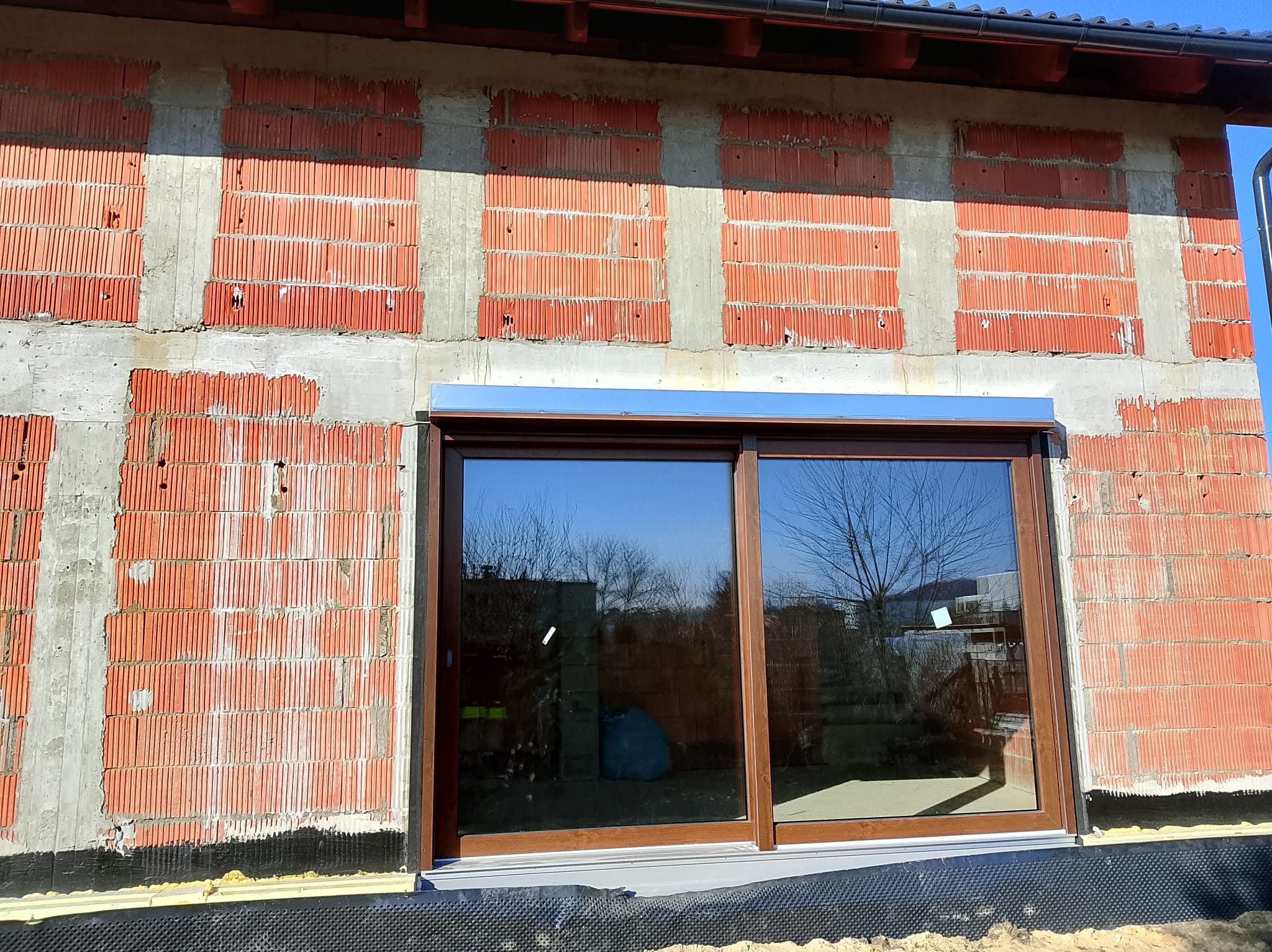 Montaż okna przesuwnego z brązową ramą w surowym murze z czerwonej cegły, widoczne odbicie drzewa w szybie.