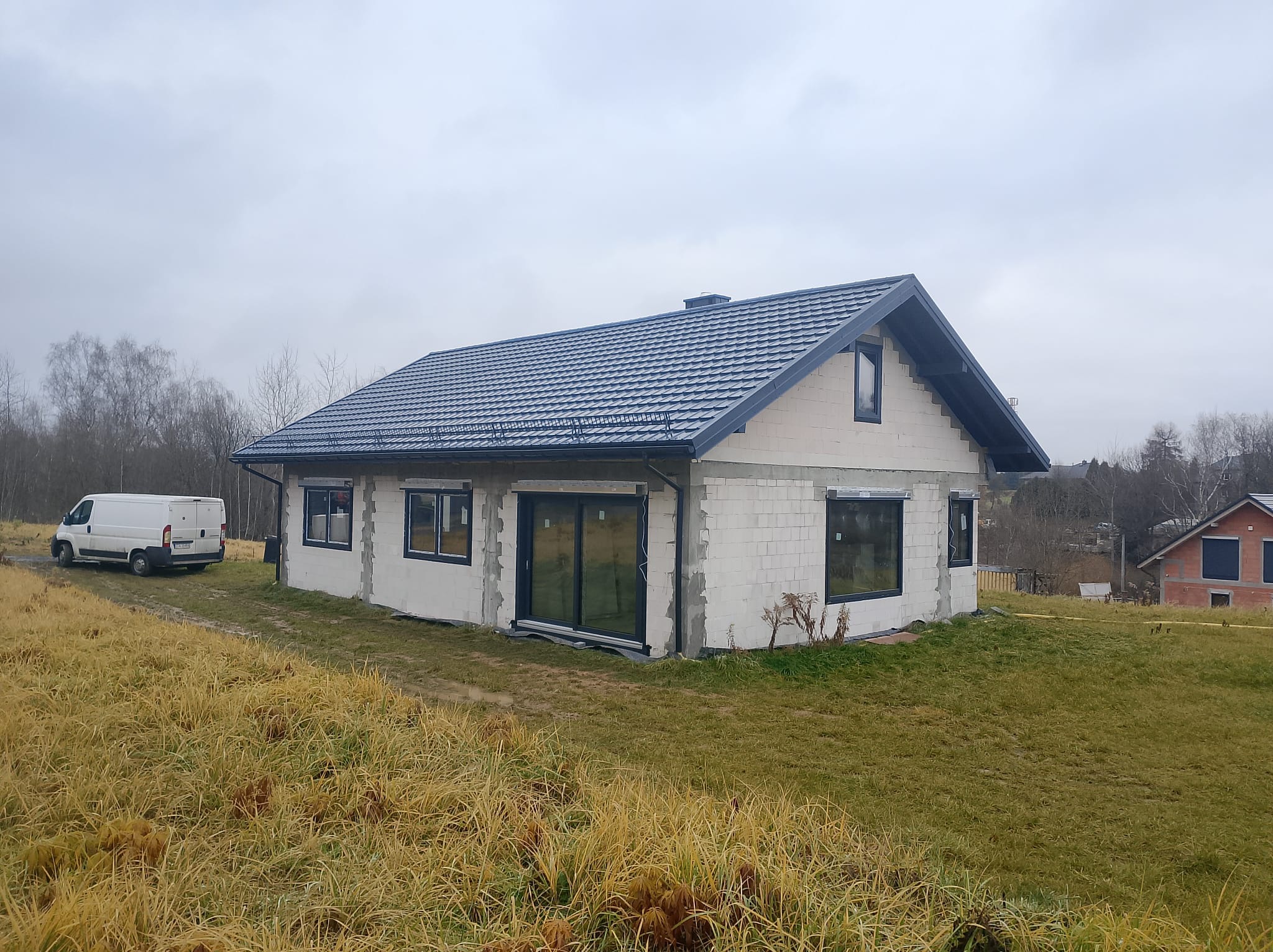 Surowy dom z białego bloczka z zamontowanymi oknami PCV i przesuwnymi drzwiami balkonowymi w czarnych ramach, na tle pochmurnego nieba i jesiennego krajobrazu, z białym busem zaparkowanym obok.