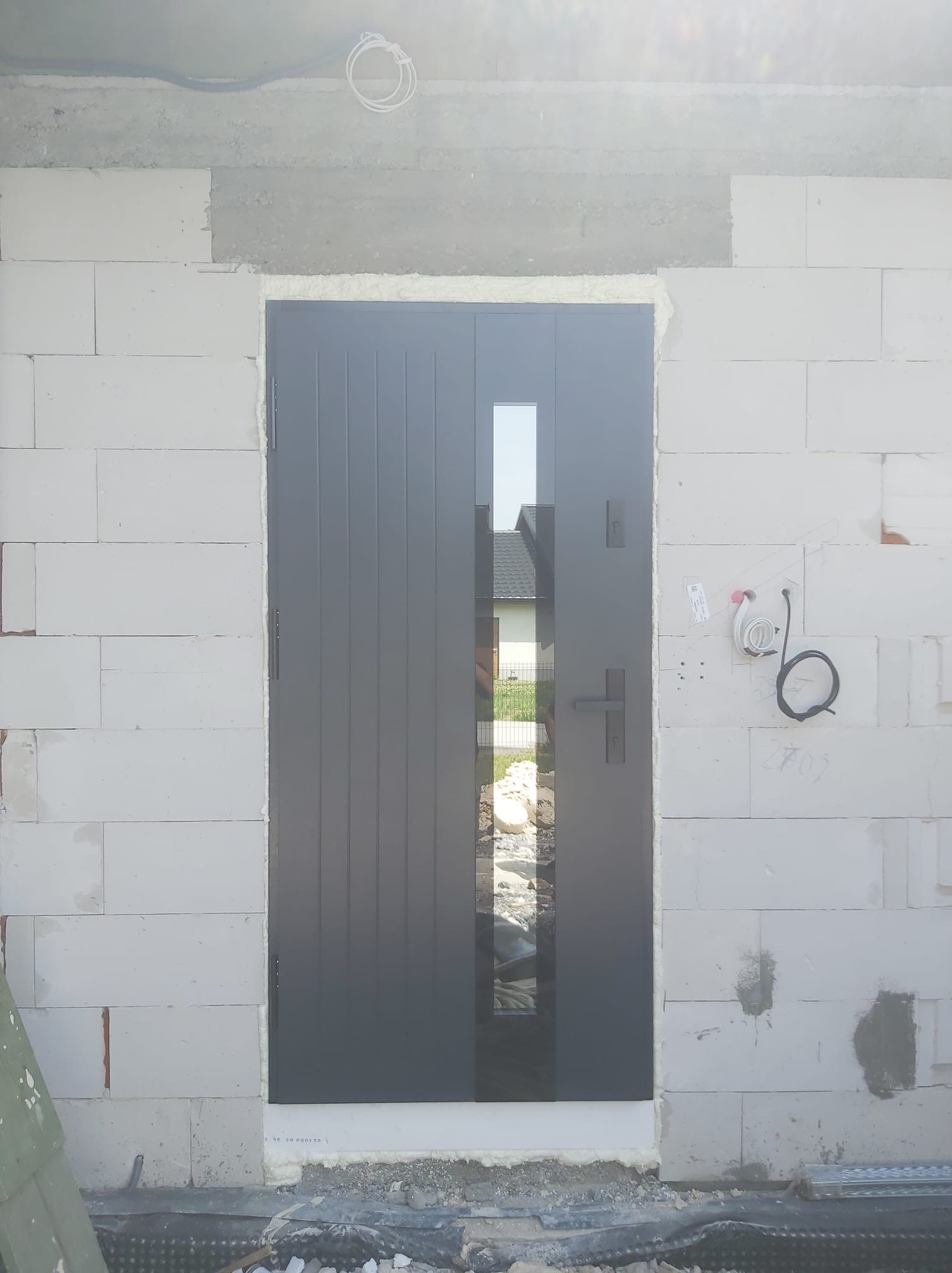 Szare drzwi zewnętrzne z pionowymi panelami i szybą, zamontowane w surowej ścianie z białych bloczków, widoczne przewody instalacji elektrycznej.