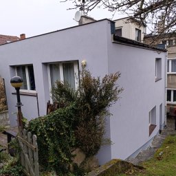 Budowa - Świeżo pomalowana na szaro elewacja domu z widoczną fakturą, zarośnięty ogródek z drewnianym płotkiem i lampą uliczną na pierwszym planie.