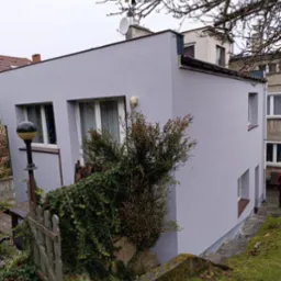 Świeżo pomalowana na szaro elewacja domu z widoczną fakturą, zarośnięty ogródek z drewnianym płotkiem i lampą uliczną na pierwszym planie.