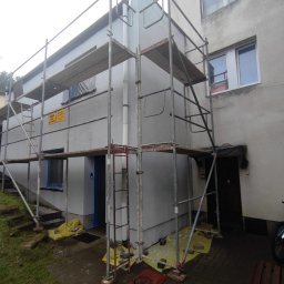 Budowa - Budynek w trakcie malowania elewacji, widoczne rusztowania, zabezpieczone folią okna i podłoże, fragment ściany już pomalowany na szaro, pozostała część budynku z nieodnowioną elewacją.