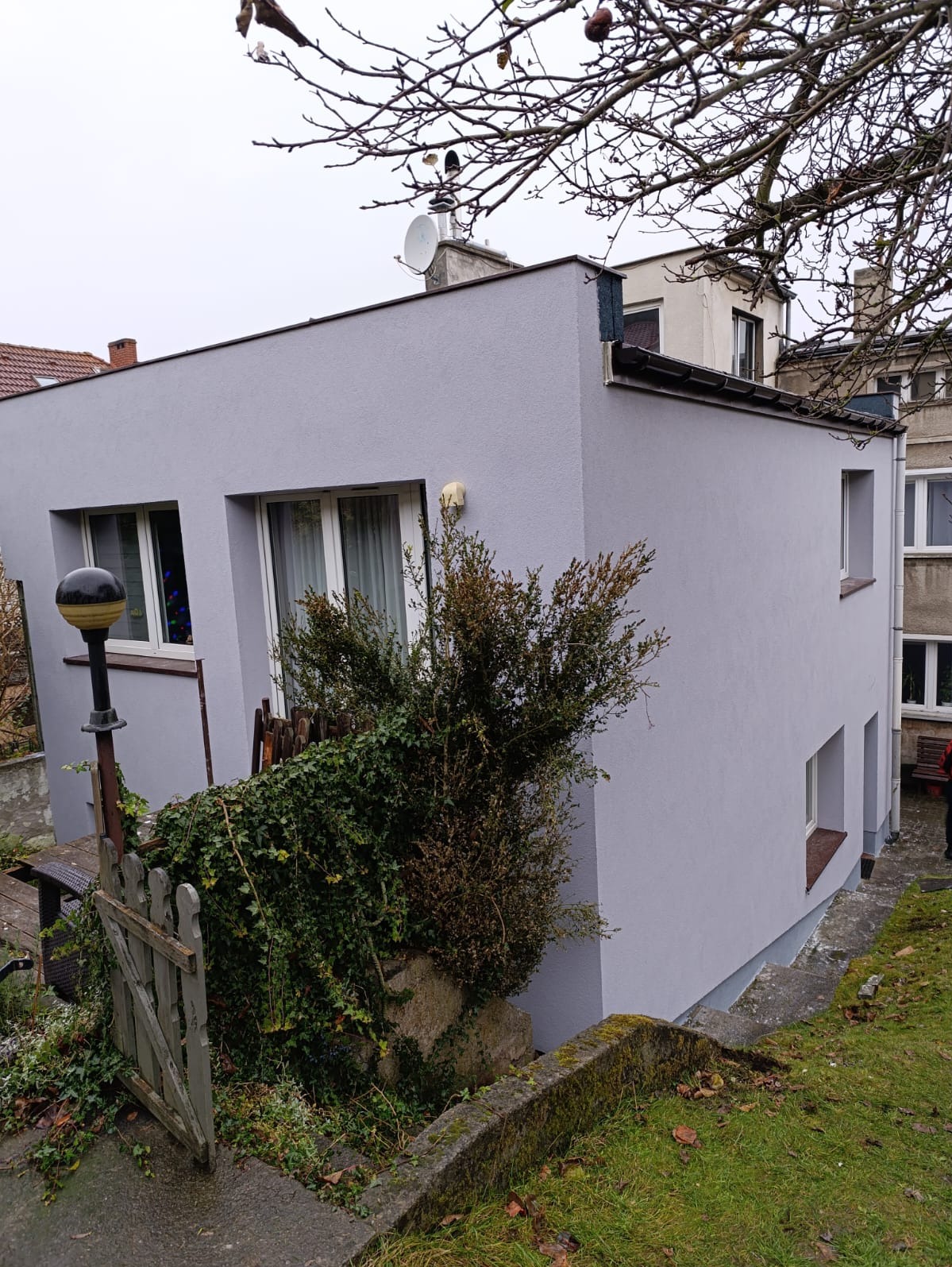 Świeżo pomalowana na szaro elewacja domu z widoczną fakturą, zarośnięty ogródek z drewnianym płotkiem i lampą uliczną na pierwszym planie.