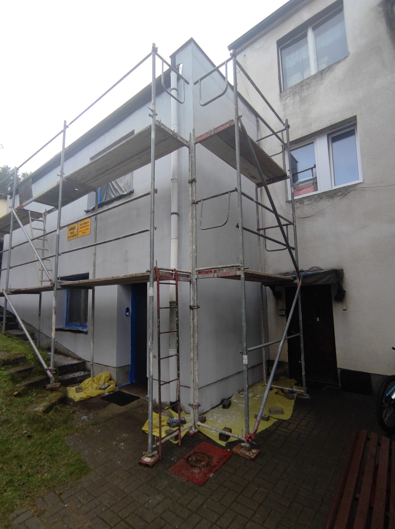 Budynek w trakcie malowania elewacji, widoczne rusztowania, zabezpieczone folią okna i podłoże, fragment ściany już pomalowany na szaro, pozostała część budynku z nieodnowioną elewacją.