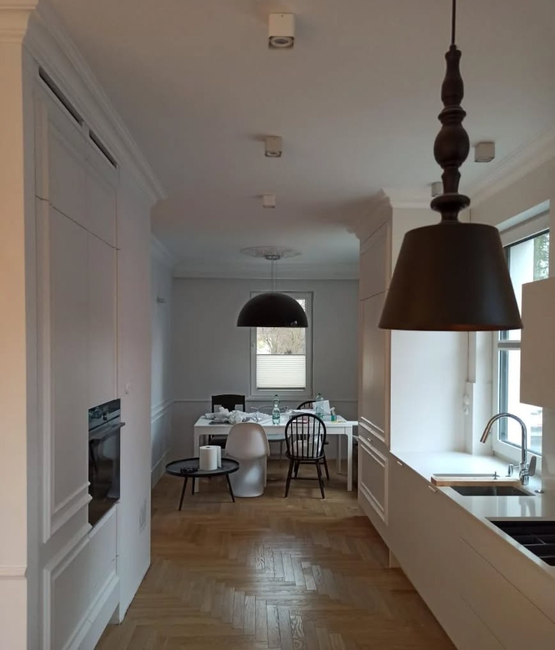 Widok na świeżo wykończone wnętrze kuchni z parkietem w jodełkę, białymi meblami z frezowanymi frontami i czarnymi lampami wiszącymi.