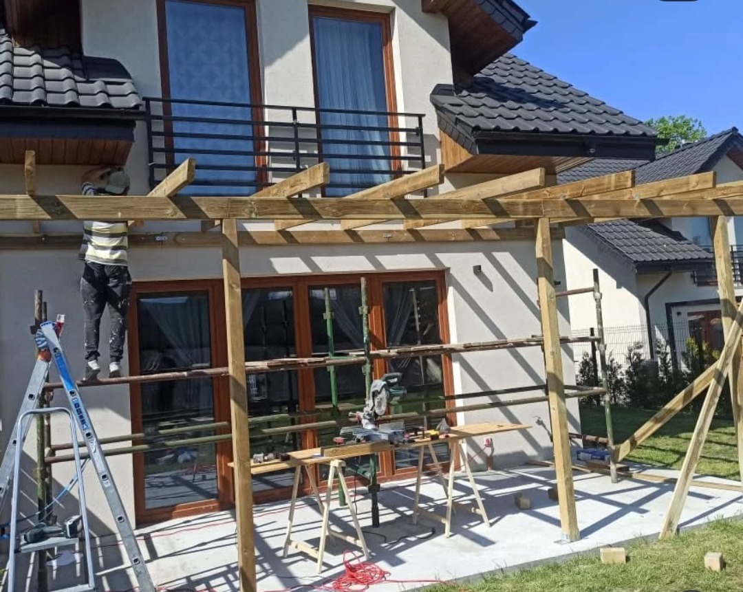 Montaż drewnianej pergoli przy domu jednorodzinnym: pracownik na drabinie łączy elementy konstrukcji, widoczne narzędzia i materiały budowlane.