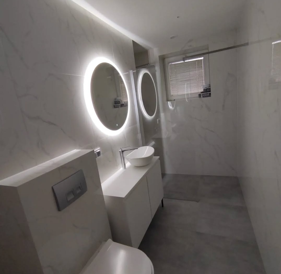 Wykończona łazienka w Gdańsku z białymi marmurkowymi płytkami, podwieszaną toaletą, umywalką na szafce i okrągłymi lustrami z podświetleniem LED.