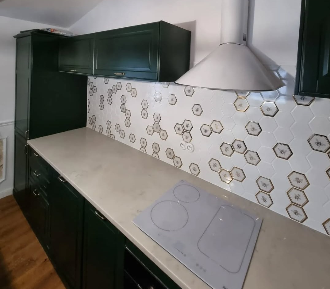 Nowoczesna kuchnia z ciemnozielonymi szafkami, blatem z jasnego konglomeratu i heksagonalnymi płytkami z motywem pszczół na ścianie, widoczna płyta indukcyjna.