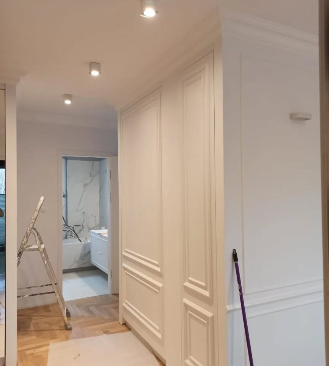 Białe, klasyczne panele ścienne w trakcie montażu, widoczna drabina i zabezpieczona podłoga parkietowa, łazienka z marmurową ścianą w tle.