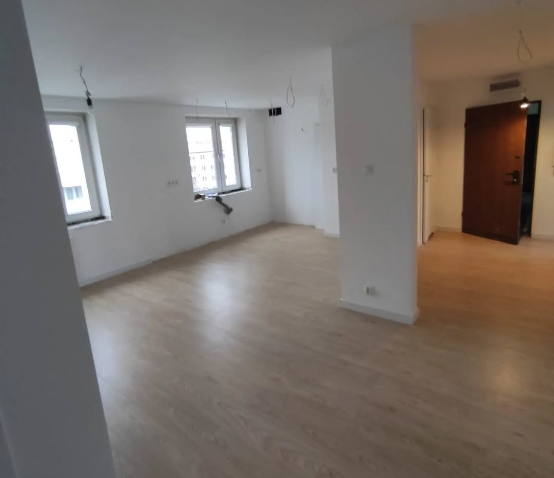 Puste, jasne pomieszczenie z parkietem, białymi ścianami i dwoma oknami, widoczne kable wystające ze ściany, drzwi wejściowe w tle.