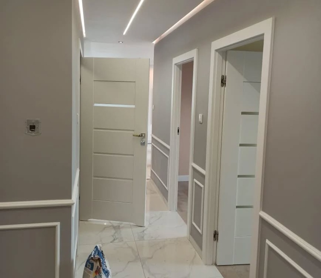 Korytarz po remoncie: szare ściany z dekoracyjnymi panelami, białe drzwi z przeszkleniami, marmurowe płytki na podłodze, oświetlenie LED w suficie.