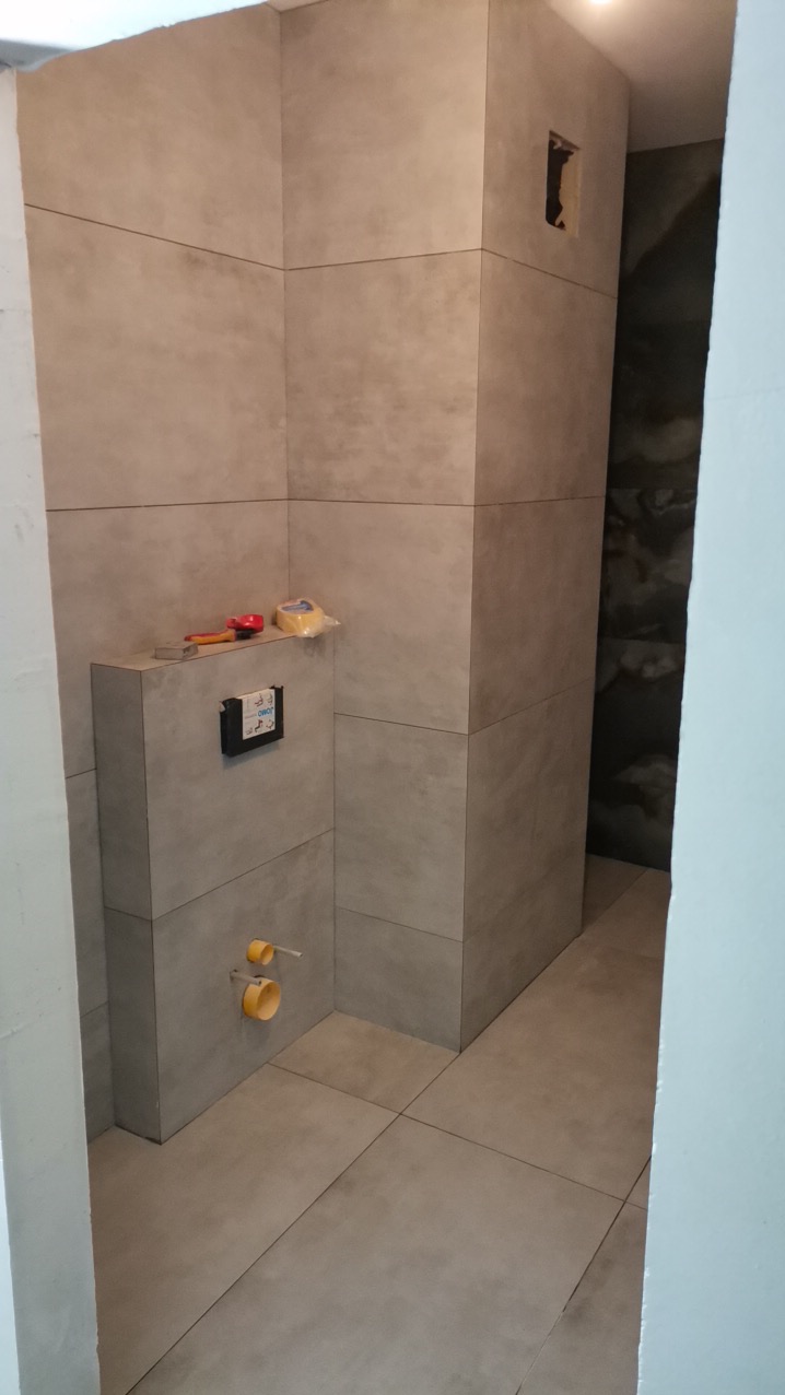 Montaż podtynkowej instalacji WC w łazience, widoczne szare płytki imitujące beton, wystające rury kanalizacyjne i narzędzia na obudowie stelaża.