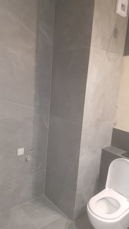 Wykończona łazienka z szarymi, matowymi płytkami imitującymi kamień, widoczny fragment toalety, włącznik światła i przyłącze wodne.
