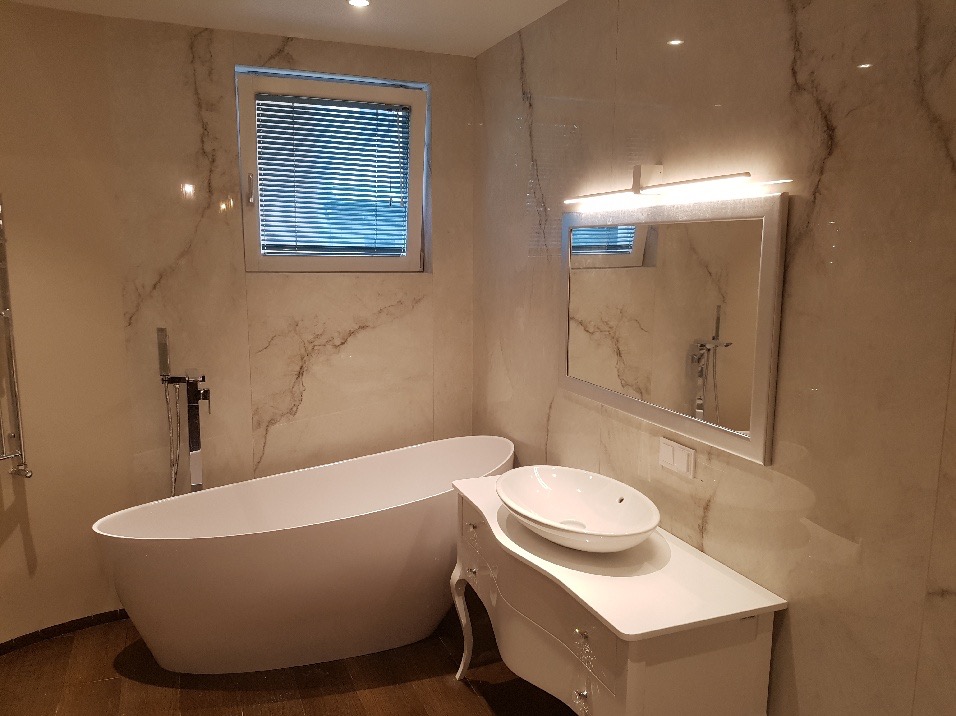 Elegancka łazienka w beżowych marmurowych płytkach, z wolnostojącą wanną, umywalką nablatową na białej szafce w stylu retro i lustrem z oświetleniem LED.