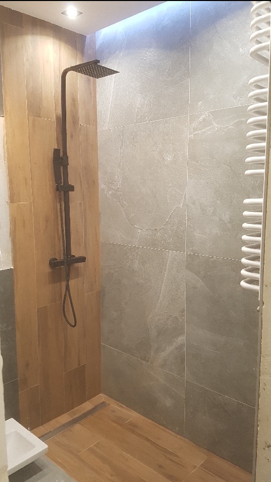 Nowoczesna kabina prysznicowa walk-in z czarną armaturą, drewnopodobnymi płytkami i szarą glazurą imitującą kamień, oświetlona punktowo i listwą LED.